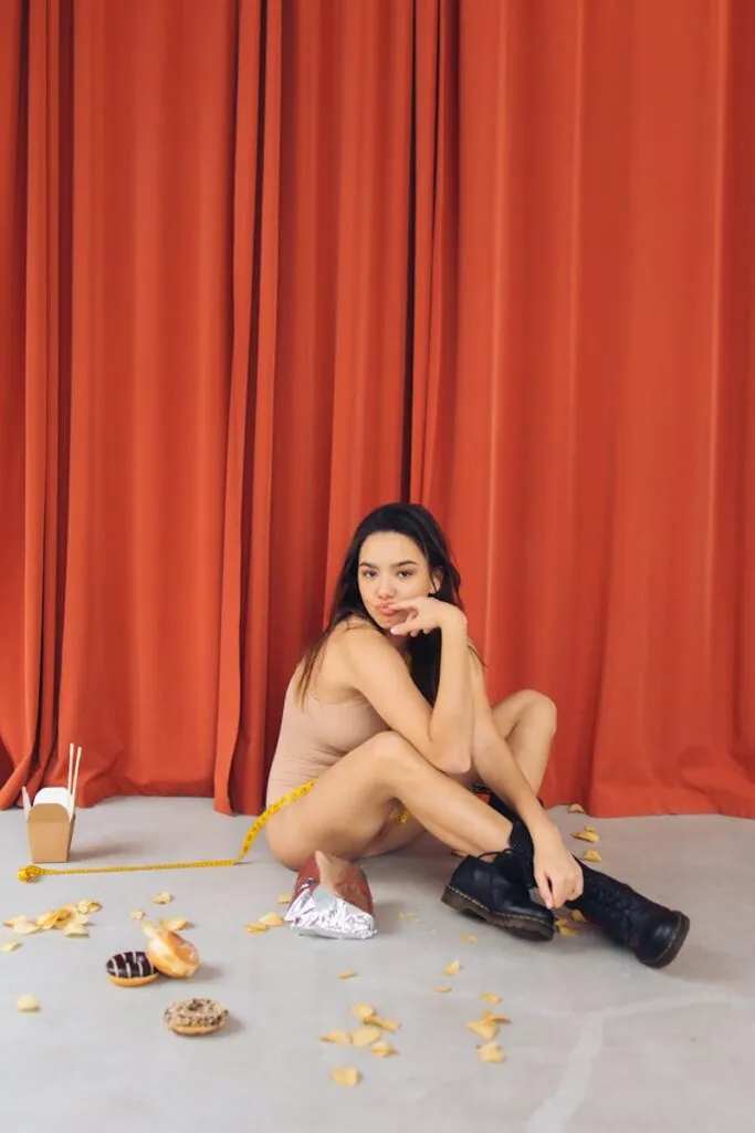 A woman sits on the floor surrounded by snacks, pondering with a tape measure beside her.