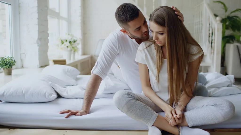 A man and woman sitting on a bed in a cozy and comforting indoor setting.