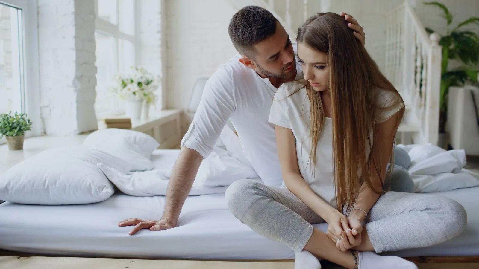 A man and woman sitting on a bed in a cozy and comforting indoor setting.